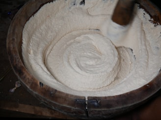 A large wooden bowl is filled with a thick, creamy mixture being stirred. The creamy substance is smooth and has a white, fluffy texture. A wooden stirring implement is visible, indicating the mixture is being manually mixed or pounded.