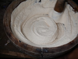 A large wooden bowl is filled with a thick, creamy mixture being stirred. The creamy substance is smooth and has a white, fluffy texture. A wooden stirring implement is visible, indicating the mixture is being manually mixed or pounded.