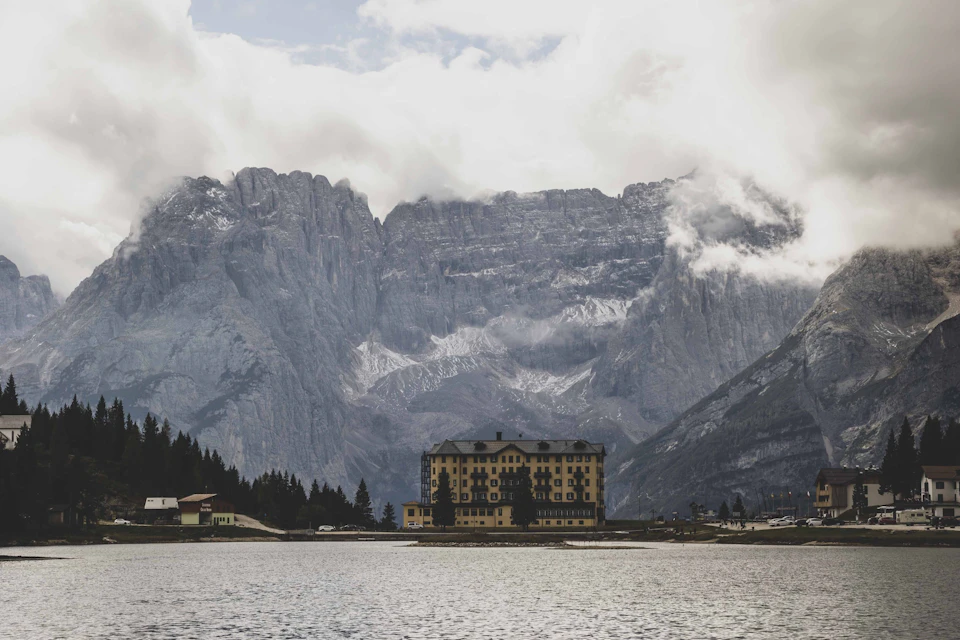 The image depicts a serene mountain landscape with a large, yellow building nestled among snow-capped mountains and surrounded by trees. The sky above is filled with clouds, casting a soft light over the scene. In the foreground, there's a body of water that reflects the surrounding scenery, adding depth to the image.