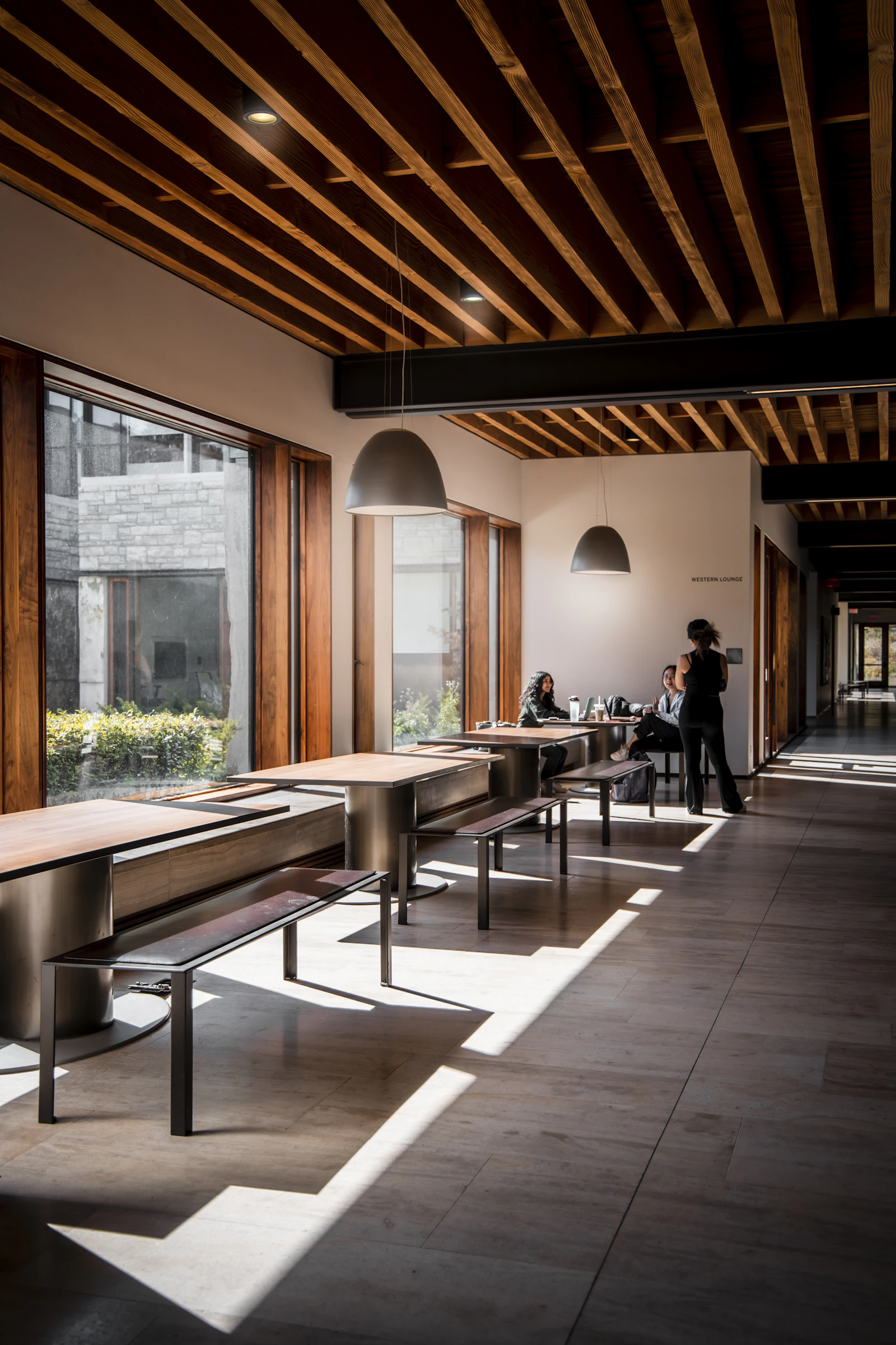 Students working at long tables and benches beside a large window in a business school building