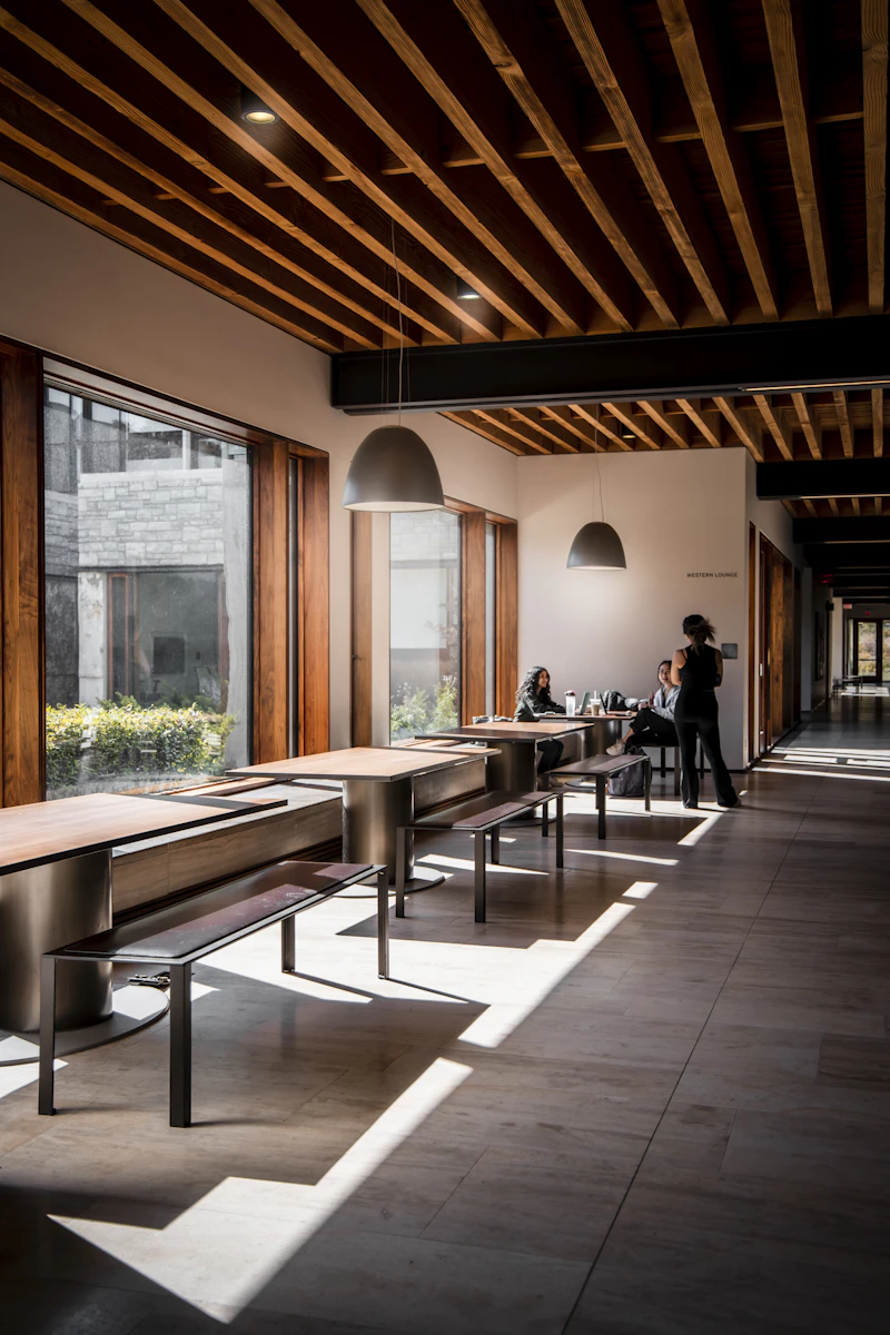 Students working at long tables and benches beside a large window in a business school building