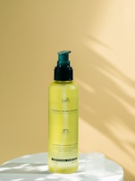 A yellow bottle of hair care product with a green pump sits on a white surface. The background is a smooth, pale yellow surface with soft shadows.