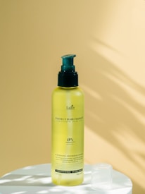 A yellow bottle of hair care product with a green pump sits on a white surface. The background is a smooth, pale yellow surface with soft shadows.