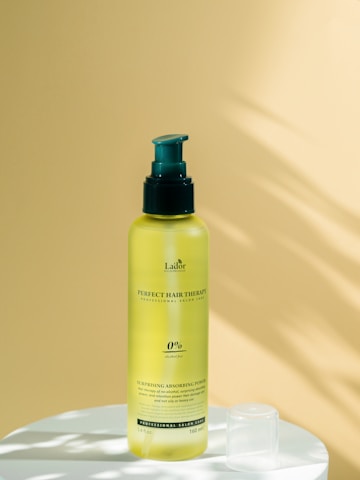 A yellow bottle of hair care product with a green pump sits on a white surface. The background is a smooth, pale yellow surface with soft shadows.