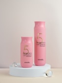 Two pink bottles of Probiotics Color Radiance Shampoo with the brand name MASIL are displayed on a white circular platform. They are accompanied by three translucent spheres, one of which is slightly larger.
