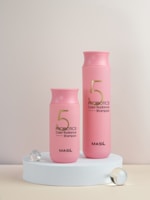 Two pink bottles of Probiotics Color Radiance Shampoo with the brand name MASIL are displayed on a white circular platform. They are accompanied by three translucent spheres, one of which is slightly larger.