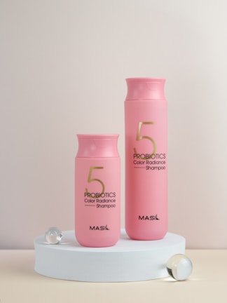 Two pink bottles of Probiotics Color Radiance Shampoo with the brand name MASIL are displayed on a white circular platform. They are accompanied by three translucent spheres, one of which is slightly larger.