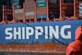 A large cargo ship is prominently displayed with the word 'SHIPPING' painted in bold white letters on its blue hull. The upper section of the ship is stacked with multicolored shipping containers. A person dressed in an orange jacket is in the foreground, partially visible, facing towards the ship.