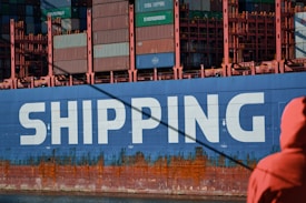 A large cargo ship is prominently displayed with the word 'SHIPPING' painted in bold white letters on its blue hull. The upper section of the ship is stacked with multicolored shipping containers. A person dressed in an orange jacket is in the foreground, partially visible, facing towards the ship.