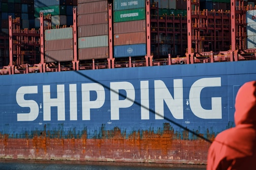 A large cargo ship is prominently displayed with the word 'SHIPPING' painted in bold white letters on its blue hull. The upper section of the ship is stacked with multicolored shipping containers. A person dressed in an orange jacket is in the foreground, partially visible, facing towards the ship.