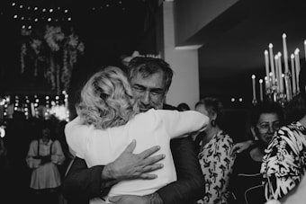 A black and white photo captures two people sharing a heartfelt embrace in a crowded, warmly lit setting. The scene suggests an emotional, possibly celebratory event, with small lights decorating the background, giving an intimate atmosphere.