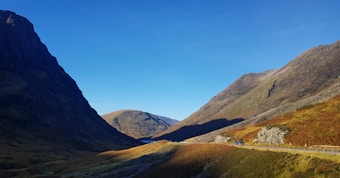 A scenic landscape featuring a wide valley surrounded by towering mountains under a clear blue sky. The sunlight casts shadows across the slopes, highlighting the contrast between light and dark areas. A road lined with guardrails curves along one side of the valley, with a vehicle visible in the distance.