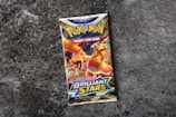 A Pokémon trading card game booster pack titled 'Sword & Shield: Brilliant Stars' is centered on a rough, textured gray surface. The pack features colorful artwork with a prominent orange and yellow dragon-like creature, exuding a dynamic and energetic feel.