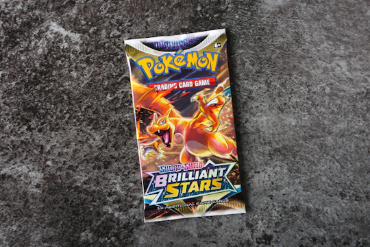 A Pokémon trading card game booster pack titled 'Sword & Shield: Brilliant Stars' is centered on a rough, textured gray surface. The pack features colorful artwork with a prominent orange and yellow dragon-like creature, exuding a dynamic and energetic feel.