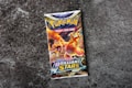 A Pokémon trading card game booster pack titled 'Sword & Shield: Brilliant Stars' is centered on a rough, textured gray surface. The pack features colorful artwork with a prominent orange and yellow dragon-like creature, exuding a dynamic and energetic feel.