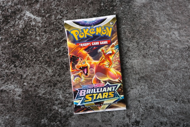 A Pok&eacute;mon trading card game booster pack titled 'Sword & Shield: Brilliant Stars' is centered on a rough, textured gray surface. The pack features colorful artwork with a prominent orange and yellow dragon-like creature, exuding a dynamic and energetic feel.