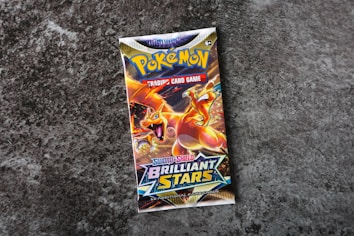 A Pok&eacute;mon trading card game booster pack titled 'Sword & Shield: Brilliant Stars' is centered on a rough, textured gray surface. The pack features colorful artwork with a prominent orange and yellow dragon-like creature, exuding a dynamic and energetic feel.