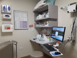 A medical examination room with a computer on a desk, storage shelves filled with medical supplies, and a BMI chart on the wall. The desk contains various bottles, papers, and electronic equipment. The room is equipped with medical gloves and a sharps disposal container mounted on the wall.