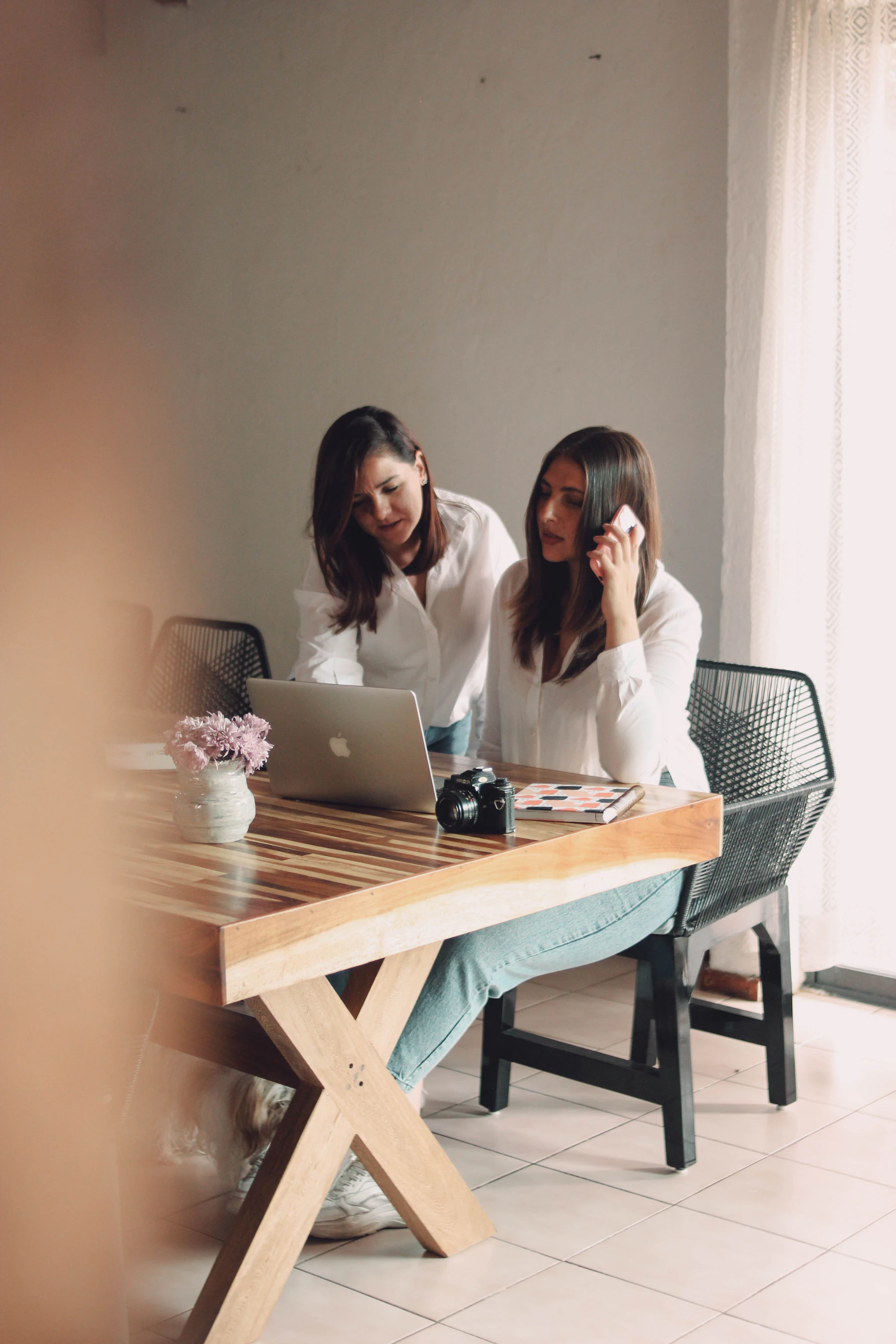 Photograph showing two people sit at a wooden table with a laptop, notebook and camera while discussing something together.