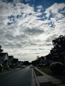 A quiet suburban street under a partly cloudy sky. The street is lined with houses featuring front lawns and driveways. Trees and bushes are scattered along the sidewalk, and a few cars are parked on the side of the road. The atmosphere suggests a peaceful residential area.