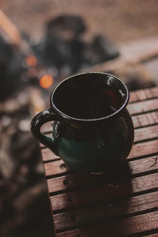Rustic enamel mugs resting on a worn wooden table beside glowing embers of a campfire.