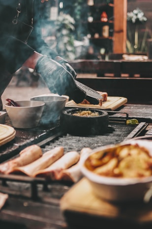 A chef wears black gloves and uses a grater over a small pot on a table filled with various food ingredients and cooking utensils. There are bowls, a cutting board, uncooked meat, and a grill, with faint smoke visible, suggesting an outdoor cooking or barbecue setup.