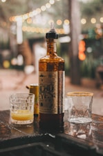 A dimly lit outdoor setting with warm, blurred lights in the background. In the foreground, an amber-colored bottle with a dropper top is flanked by two glasses. One glass contains a yellow liquid, while the other has a salted rim and is empty.