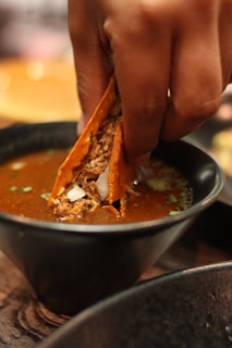 A close-up of a person's hand dipping a taco into a bowl of rich, flavorful broth. The taco is filled with shredded meat and garnished with chopped onions and herbs. The scene suggests a warm and savory dining experience.