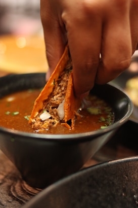 A close-up of a person's hand dipping a taco into a bowl of rich, flavorful broth. The taco is filled with shredded meat and garnished with chopped onions and herbs. The scene suggests a warm and savory dining experience.