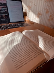 An open book lies on a wooden table, with sunlight casting shadows across the pages. In the background, a laptop is open displaying a webpage with text and images. The setting appears to be a cozy indoor workspace.