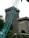 The image depicts a construction site in an urban area, featuring a large crane in the foreground. Behind the crane, there are two tall buildings with the name 'mandiri' on them. The buildings are surrounded by some trees and greenery. The sky appears overcast.