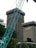 The image depicts a construction site in an urban area, featuring a large crane in the foreground. Behind the crane, there are two tall buildings with the name 'mandiri' on them. The buildings are surrounded by some trees and greenery. The sky appears overcast.
