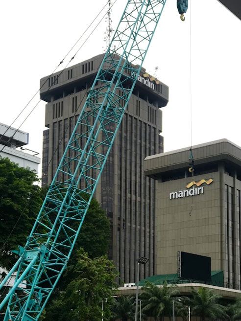 The image depicts a construction site in an urban area, featuring a large crane in the foreground. Behind the crane, there are two tall buildings with the name 'mandiri' on them. The buildings are surrounded by some trees and greenery. The sky appears overcast.