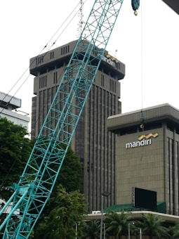 The image depicts a construction site in an urban area, featuring a large crane in the foreground. Behind the crane, there are two tall buildings with the name 'mandiri' on them. The buildings are surrounded by some trees and greenery. The sky appears overcast.