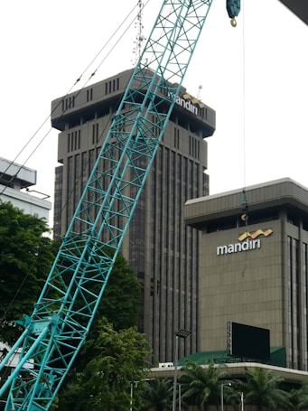 The image depicts a construction site in an urban area, featuring a large crane in the foreground. Behind the crane, there are two tall buildings with the name 'mandiri' on them. The buildings are surrounded by some trees and greenery. The sky appears overcast.