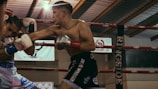 A boxing match is taking place with two boxers engaged in action inside a ring. One boxer is landing a punch as the other defends. The setting includes visible boxing gloves, ropes of the ring, and banners in the background.