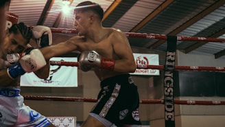A boxing match is taking place with two boxers engaged in action inside a ring. One boxer is landing a punch as the other defends. The setting includes visible boxing gloves, ropes of the ring, and banners in the background.