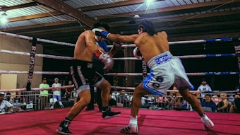 Two boxers are engaged in a match inside a boxing ring surrounded by spectators. The boxer on the right is throwing a punch at his opponent, who is dressed in black shorts and black boxing shoes. The surroundings include a crowd of people watching the fight, the boxing ring ropes, and the roof structure of the venue.
