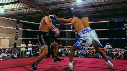 Two boxers are engaged in a match inside a boxing ring surrounded by spectators. The boxer on the right is throwing a punch at his opponent, who is dressed in black shorts and black boxing shoes. The surroundings include a crowd of people watching the fight, the boxing ring ropes, and the roof structure of the venue.