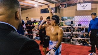 Two boxers stand in a boxing ring, with one raising his hand victoriously. A judge in a blue shirt is present, along with spectators surrounding the ring. The atmosphere suggests a recently concluded match with the winning boxer being acknowledged.