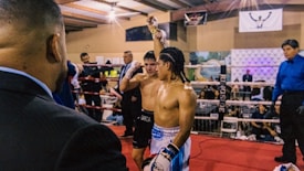 Two boxers stand in a boxing ring, with one raising his hand victoriously. A judge in a blue shirt is present, along with spectators surrounding the ring. The atmosphere suggests a recently concluded match with the winning boxer being acknowledged.