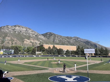 A baseball game is taking place on a well-maintained field, surrounded by a scenic mountain range under a clear blue sky. Several players are visible on the field, with one at bat and another near the pitcher's mound. The backdrop features lush green trees and a building. Ads and signs from various sponsors are displayed around the field.