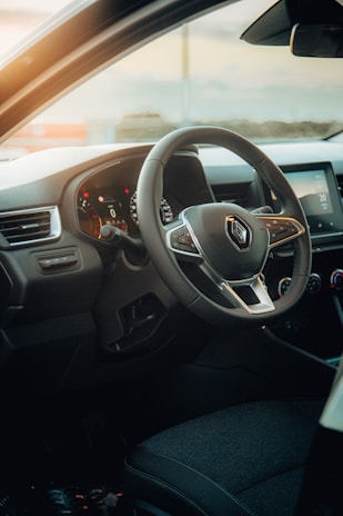 A car interior featuring a steering wheel with a Renault logo, dashboard display with illuminated gauges, and part of the driver's seat. The lighting suggests it is either early morning or late afternoon, as warm sunlight enters through the window.