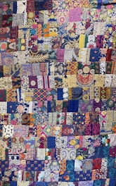 A vibrant and intricately designed quilt featuring a patchwork of various floral and abstract patterns. The patchworks include a diverse mixture of colors, with a predominant use of blues, purples, and golds. A variety of flowers are intricately woven throughout, along with paisley and geometric designs that add complexity and depth to the overall piece.