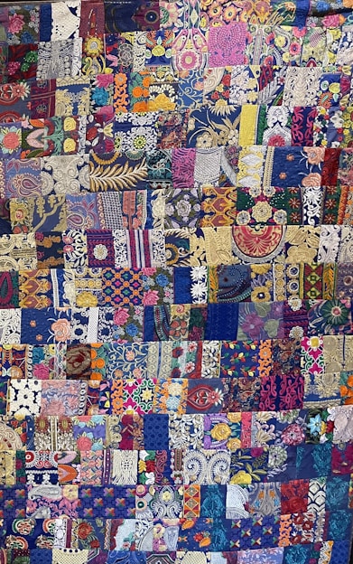 A vibrant and intricately designed quilt featuring a patchwork of various floral and abstract patterns. The patchworks include a diverse mixture of colors, with a predominant use of blues, purples, and golds. A variety of flowers are intricately woven throughout, along with paisley and geometric designs that add complexity and depth to the overall piece.