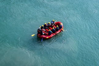 A group of people wearing helmets and life jackets are engaged in white-water rafting, paddling together in a bright red inflatable raft. The raft navigates through clear, blue-green water, suggesting an outdoor adventure or team activity.