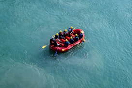 A group of people wearing helmets and life jackets are engaged in white-water rafting, paddling together in a bright red inflatable raft. The raft navigates through clear, blue-green water, suggesting an outdoor adventure or team activity.