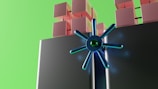 A futuristic digital illustration featuring stacked cubes in shades of pink and a large, metallic blue circular structure with radial spokes. The background is a bright green, adding a vibrant contrast.