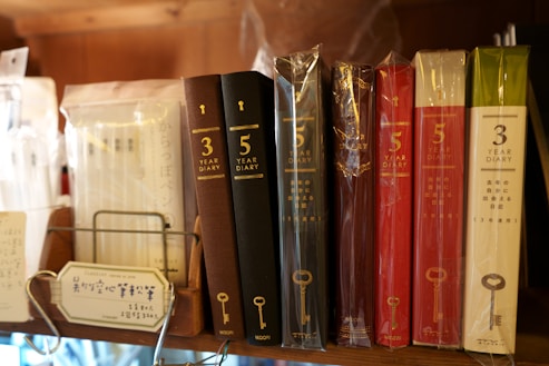 A selection of diaries displayed on a shelf, with covers of various colors including brown, red, and white. The diaries are labeled for three-year and five-year use, and some are wrapped in plastic. There are also packs of clear pens on the shelf, along with a small informational sign in Japanese.