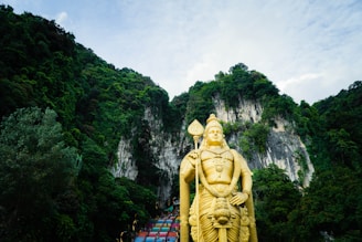 A large golden statue stands prominently against a backdrop of lush green mountains and a partly cloudy sky. The structure is intricately detailed and holds a scepter, conveying a sense of grandeur and reverence. The surrounding dense foliage creates a striking natural setting.